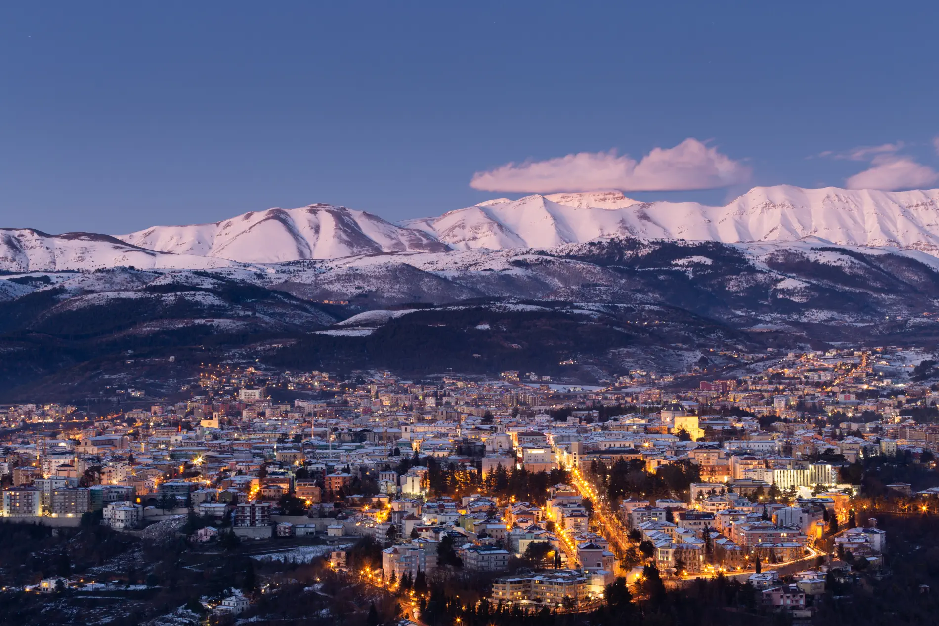 Foto con panorama della città dell'Aquila e le montagne innevate sullo sfondo durante l'ora blu
